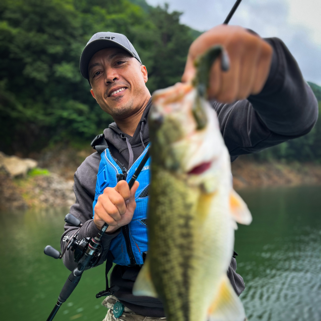 Andre proudly holding a largemouth bass he caught while fishing at a scenic green lake, wearing a blue life vest and black cap.